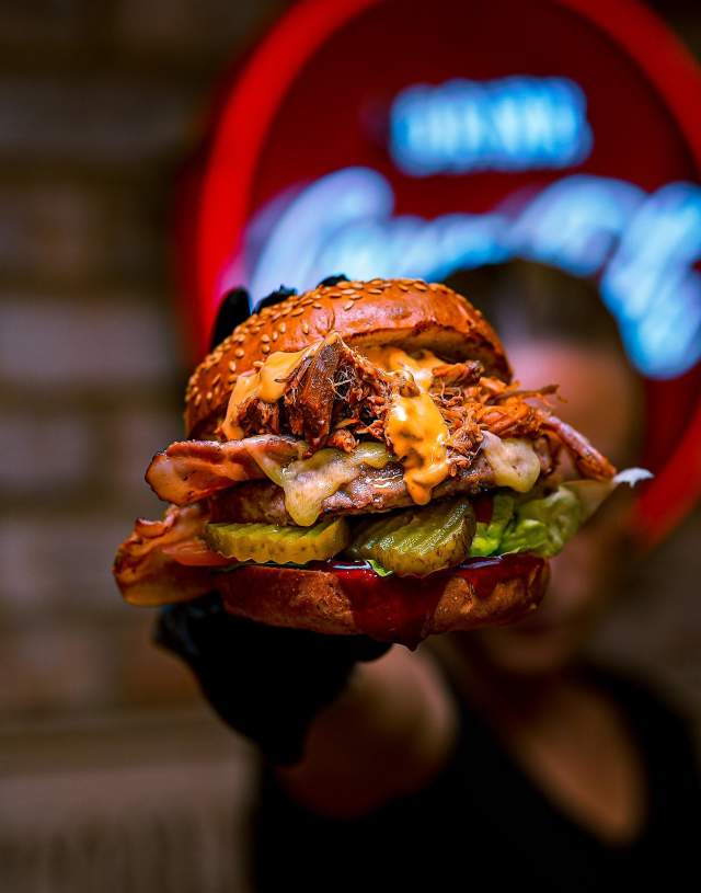A close up of a man holding a burger up to camera in a dim lit restaurant