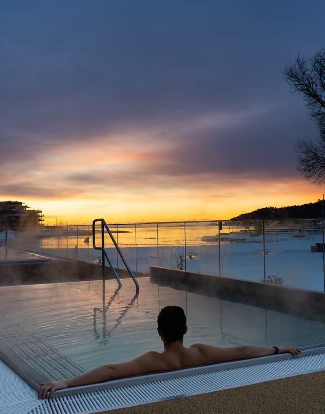 Person sitter med ryggen mot kameraet i et utendørs badebasseng i vinterlandskap