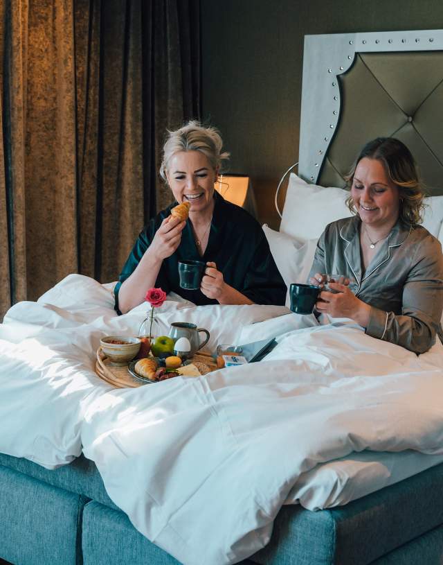 To damer som spise mat og drikke kaffe i en hotellseng. De ler og hygger seg.