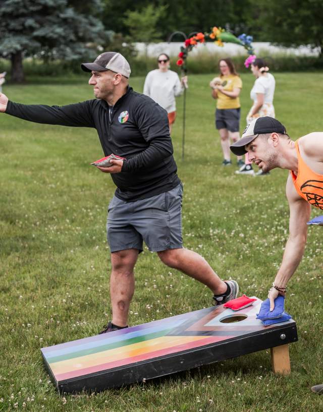 Two men playing cornhole at a Pride event in Mason. There are rainbow colors on the cornhole board and people in the background.