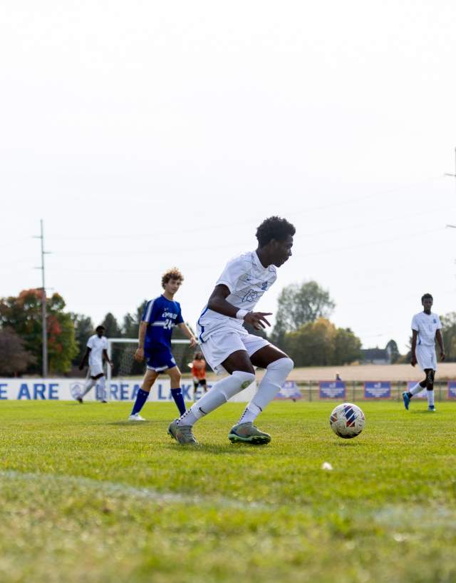 A soccer player in a white jersey and shorts, running with a soccer ball. He has dark hair and is wearing gray cleats. A player in a blue jersey is behind him, and another player is in the background in a white jersey. The ground is green turf.