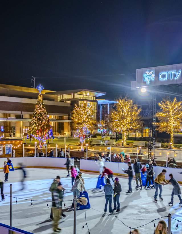 An image of an outdoor ice skating rink at City Springs. It is nighttime, the trees are illuminated with lights, and there are people on the ice skating.