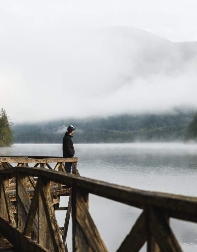 A man stands on a boardwalk looking out over the misty lake.