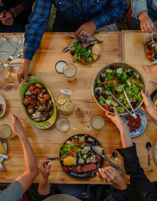 An overhead view of several people gathered around a wooden outdoor table, sharing a meal of grilled meats, corn, potatoes, and fresh salads in a warm evening setting.
