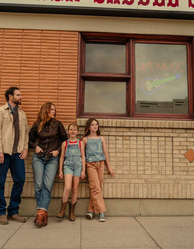 A family stands outside a downtown business in Cody, Wyoming, beneath a neon “Live Music” sign. Western-inspired clothing, cowboy boots and the historic streetscape reflect Cody’s family-friendly atmosphere, local culture and Western heritage.