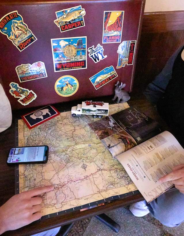 Two travelers plan a Wyoming road trip using a state map and Wyoming travel guide. Wyoming-themed stickers, a miniature truck and travel materials surround the map as one traveler points to a route, highlighting trip planning and exploration across the state.