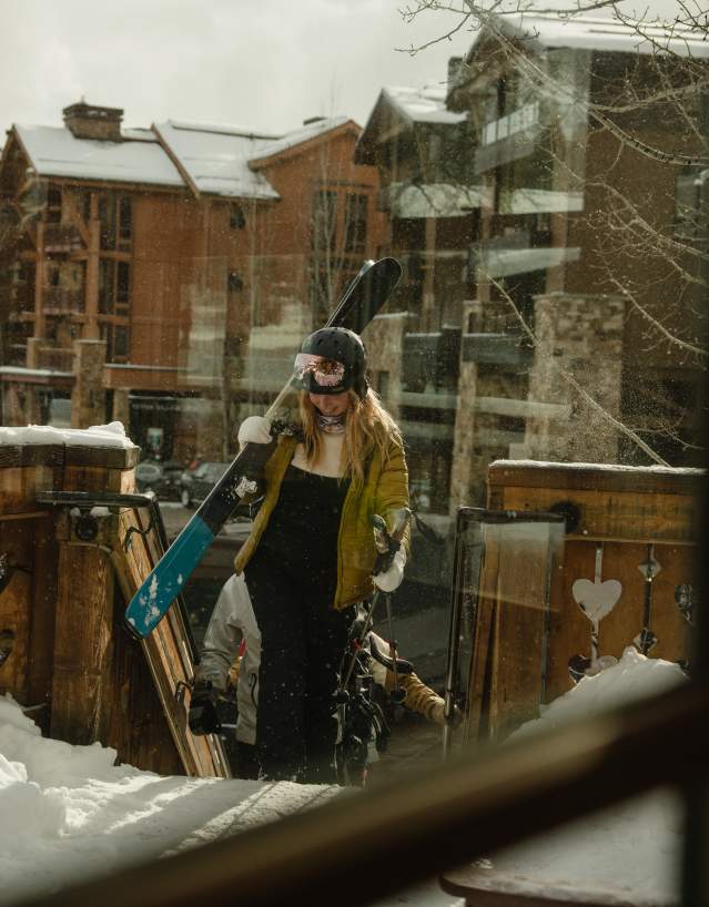 A skier carrying skis walks through deep snow toward a lodge after a day on the slopes near Jackson, Wyoming. Snow-covered buildings and a rustic wooden deck frame the winter scene, highlighting the après-ski experience and mountain lifestyle in Teton County.