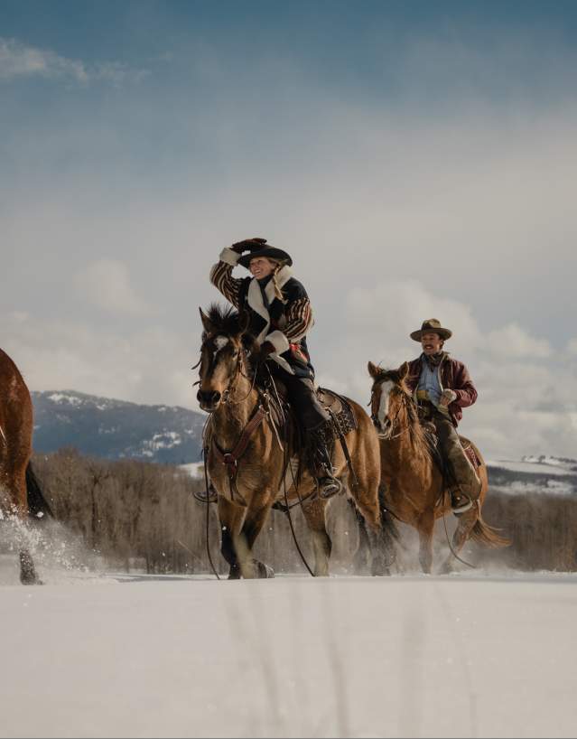 Four horseback riders travel through deep snow near Jackson, Wyoming, with forested hills and snow-covered mountains in the distance. Dressed in Western winter attire, the group rides across the open landscape, highlighting horseback riding and Wyoming’s cowboy culture during the winter season.