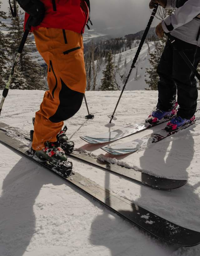 Two skiers pause on a snow-covered mountain slope near Jackson, Wyoming, with a wintry mountain landscape and evergreen trees in the background. The close-up view of skis, poles and winter gear highlights downhill skiing and outdoor recreation in Teton County.