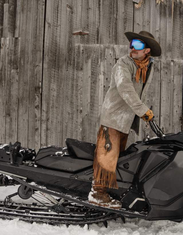 A cowboy dressed in winter Western attire stands on a snowmobile beside a weathered wooden barn near Jackson, Wyoming. Snow-covered chaps, a cowboy hat and ski goggles blend Wyoming’s Western heritage with modern winter recreation in Teton County.