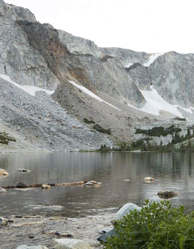 Gray rock cliff side leading down to a glacial lake with sparce pine trees surounding it with a man, a boy and a girl fishing in the lake.