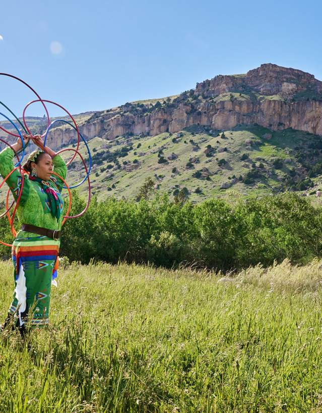 Woman dressed in regalia holding hoops overhead in prairie