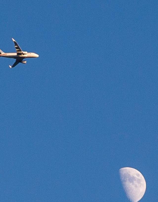 An airplane flying across a clear blue sky with the moon in view.