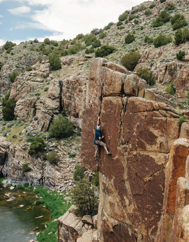 A person rock climbs a tall canyon wall above a winding river, surrounded by rugged cliffs and scattered vegetation.