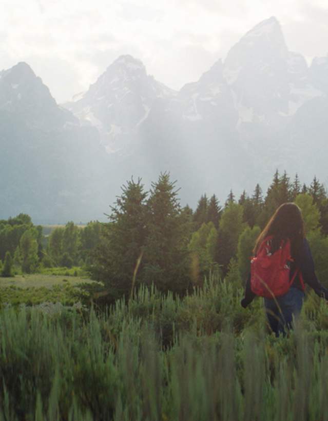Girl with backback walking through field set against a spectacular mountain view in the distance.