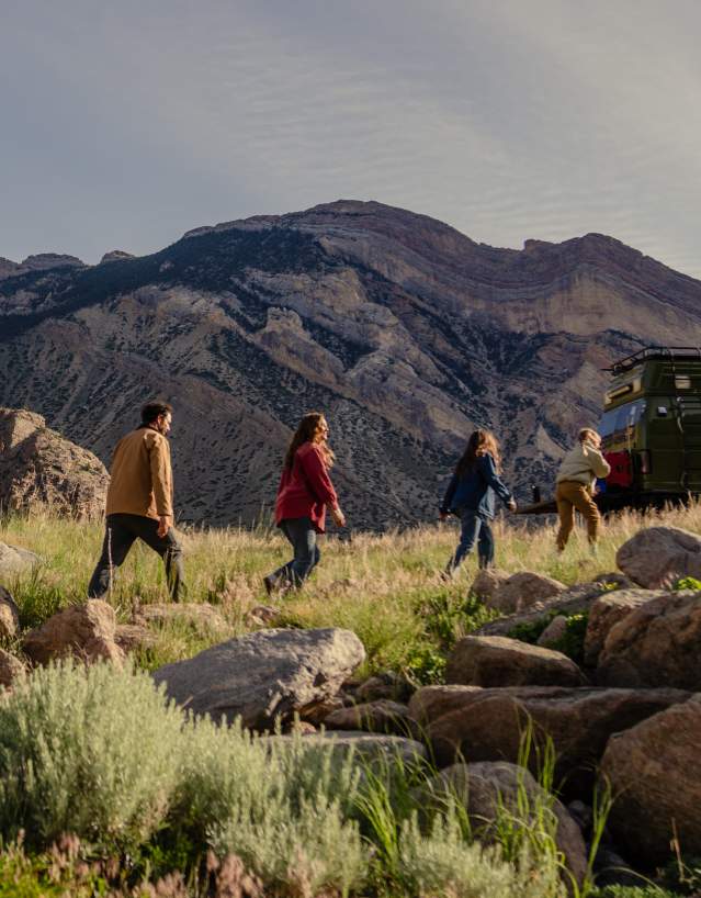 Four people walk through a grassy, rocky mountain landscape toward a parked green off-road vehicle, with steep, rugged cliffs and layered mountains rising in the background under a lightly clouded sky.