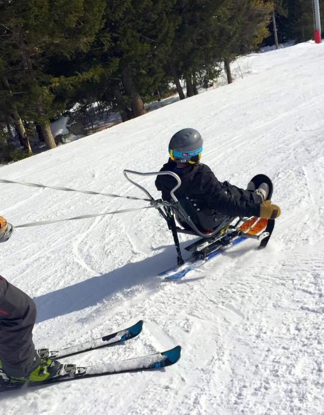 An adaptive skier uses a sit-ski while being assisted by an instructor on a snow-covered ski slope in Wyoming. The skier wears a helmet and winter gear as they navigate the mountain, highlighting accessible outdoor recreation and adaptive skiing opportunities.