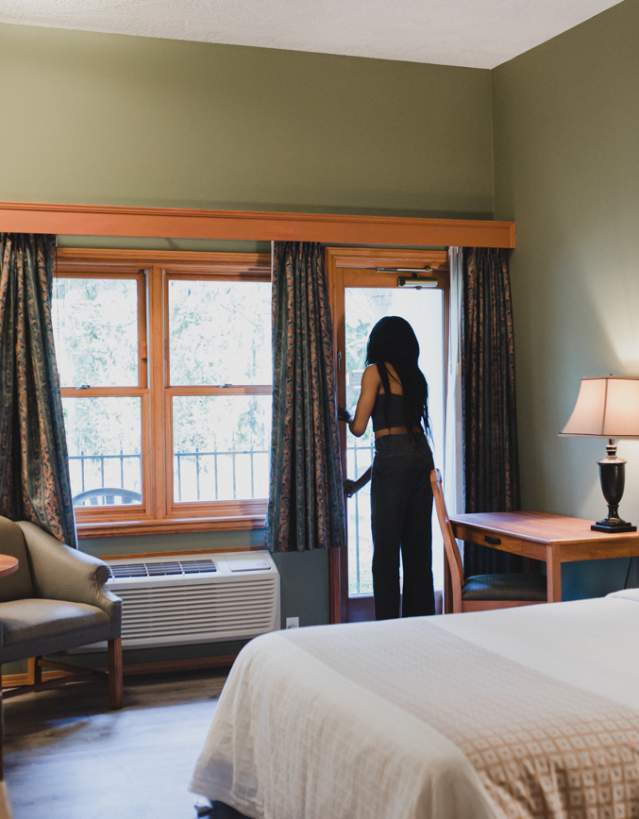 A cozy hotel room with a bed, desk, and seating area as a person opens a balcony door to let in natural light.