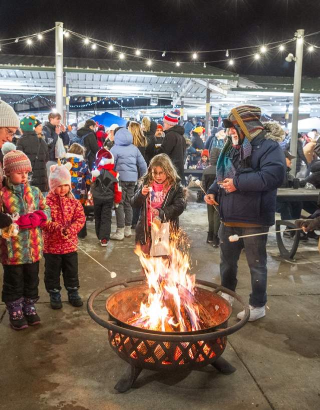 people of all ages stand outside on concrete circled around an active firepit, some children toast marshmellows on a stick over the fire as one person assists.  Others stare at the fire