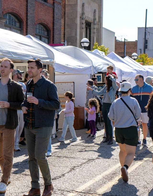 People walking around in a downtown street lined with vendor booths filled with artwork. People pass both ways, two men with coffee in their hand. Others walk in the opposite direction.