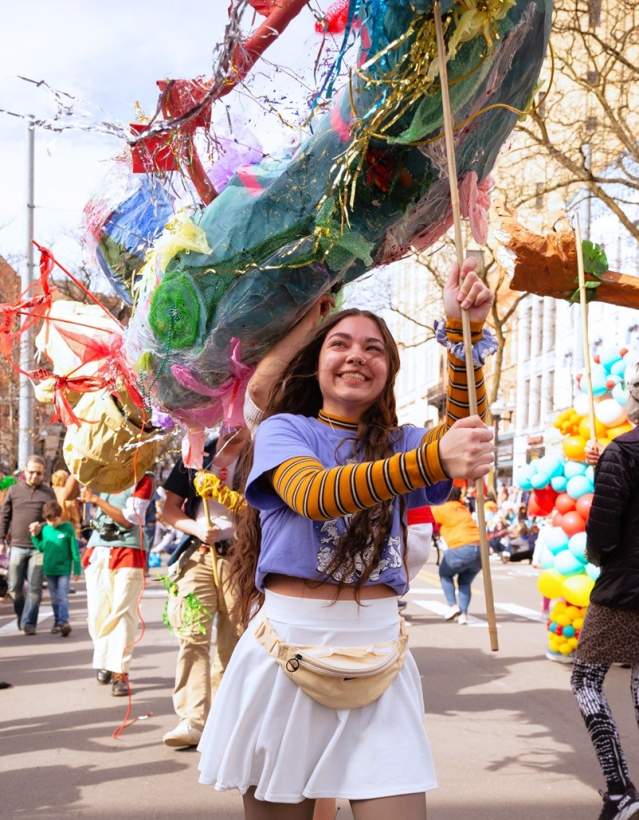 Young woman smiles as she helps support a large puppet with a wooden post during a street festival parade.