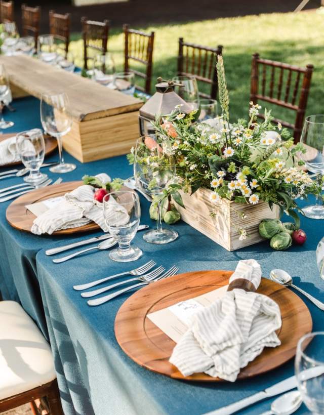 Outdoor table setting with wood and white chairs, blue tablecloth, and flowers