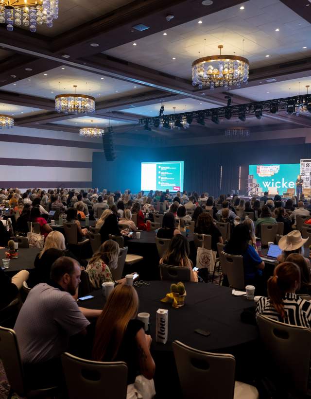 Interior of a ballroom in Fairmont Hotel with meeting attendees at round tables and a speaker on the stage at the front of the room