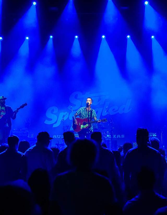 Band playing on a stage with shadows of fans in the foreground. The whole image is lit by blue lights from above.