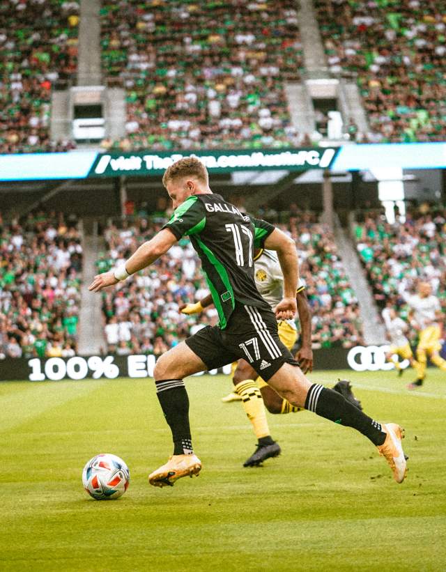 John Gallagher running with the ball at Q2 Stadium in front of a crowd.