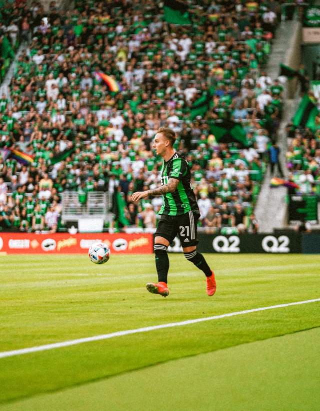 Žan Kolmanič for Austin FC on the field with the ball at Q2 stadium. A large crowd in the Supporters Section is visible behind him, waving rainbow flags