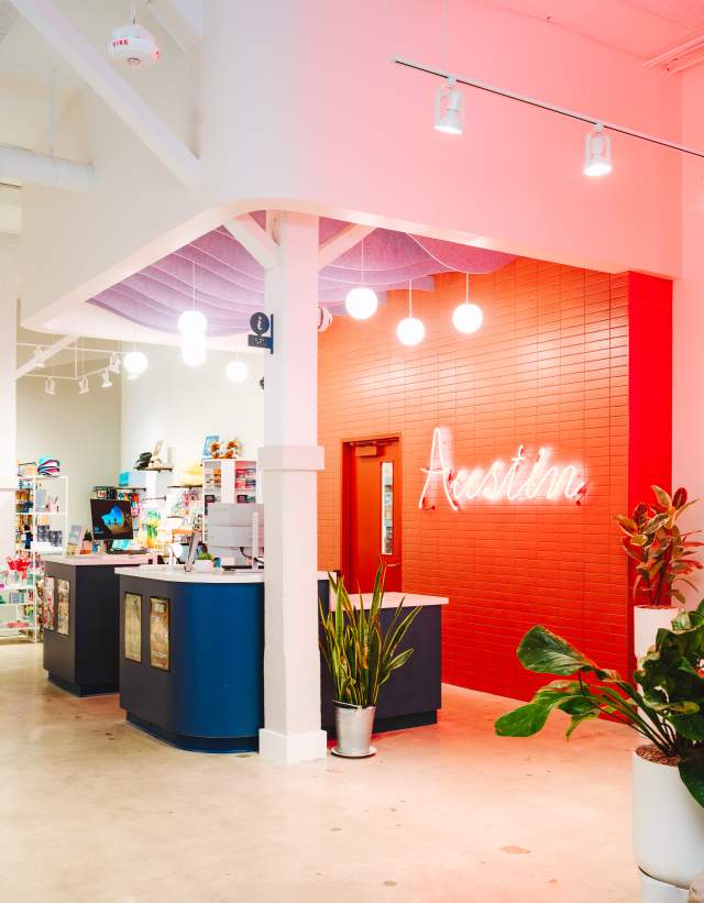 Interior of the Austin Visitor Center showing a welcome desk with brightly colored wall and "Austin" neon sign behind, and a neon Austin Motel sign with marquee reading "Jimmy Kimmel Live! Austin".
