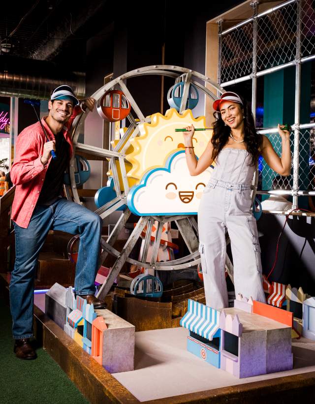 Man and woman posing at one of the holes at Holey Moley with their golf clubs and Holey Moley visors.