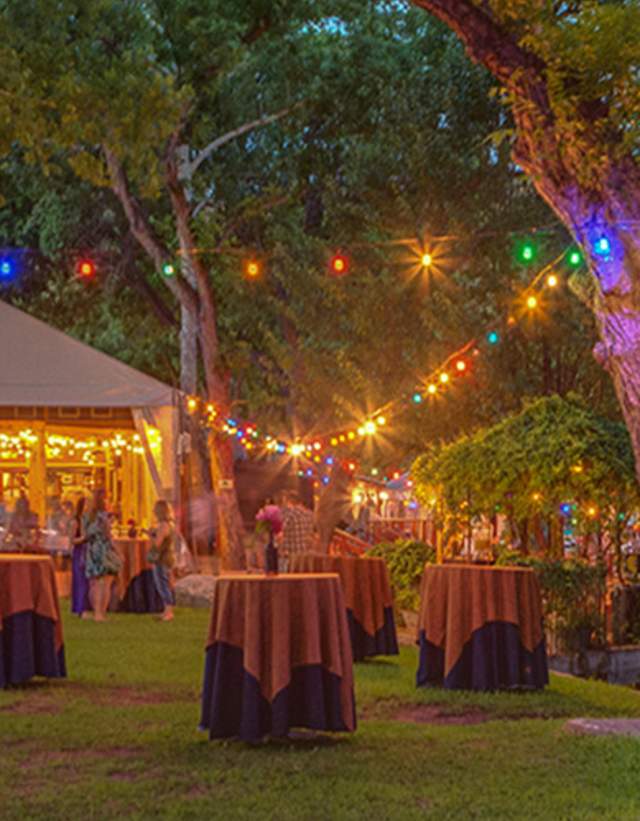 Lush green space decorated with high top tables, string lights and a large event tent.