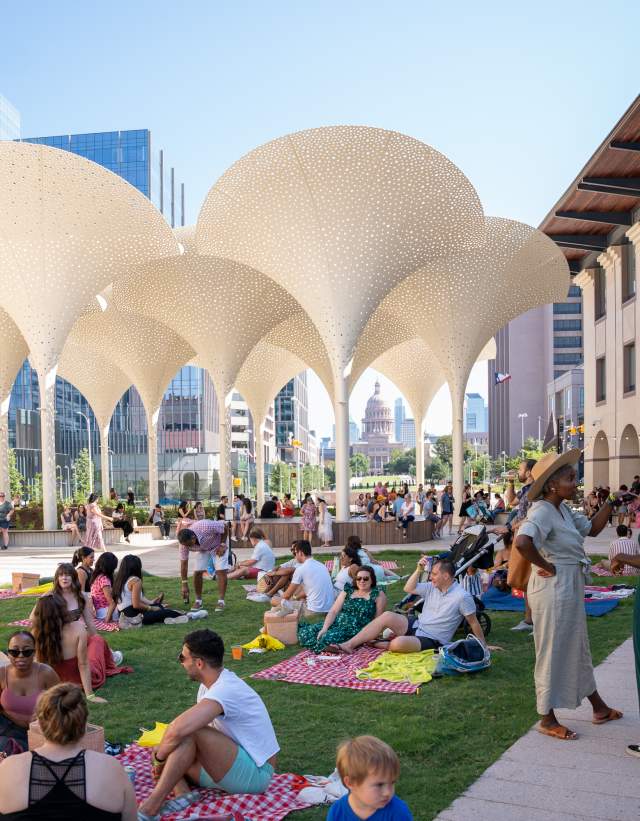 Groups of people standing around and sitting on picnic blankets across the Blanton Museum's lawn, in between museum buildings.