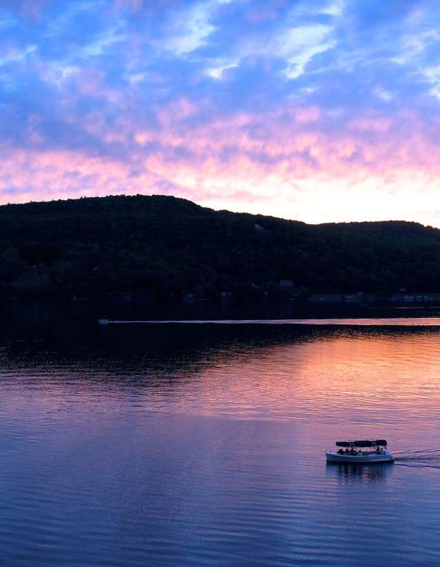 A lake with hills in the background with a boat and a blue, pink, and orange sunset in the sky and reflecting in the water.