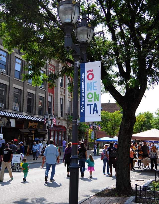 A street festival with tents, people walking through the street and a light pole with a sign reading "Oneonta" in the foreground.