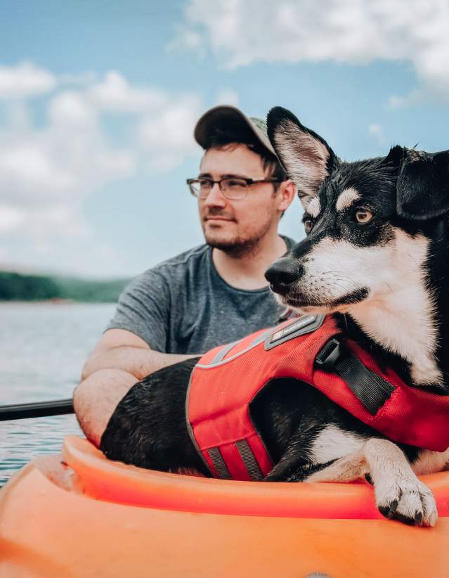 A person and a dog wearing a red life jacket in an orange kayak in a body of water with hills in the background.