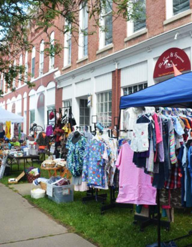 Main street with vendor tents selling items along the sidewalk with green grass in front of brick buildings.
