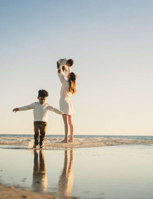 a beach at sunset with a family. one child is splashing in a tide pool, the other one is being lifted in the air by the mother