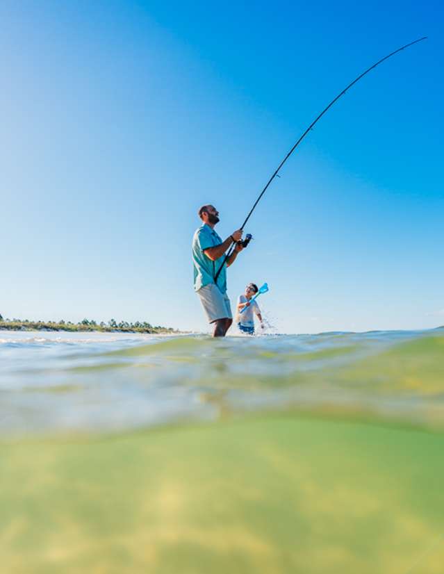 A man is fishing from the shore as his son plays in the waters beside him.