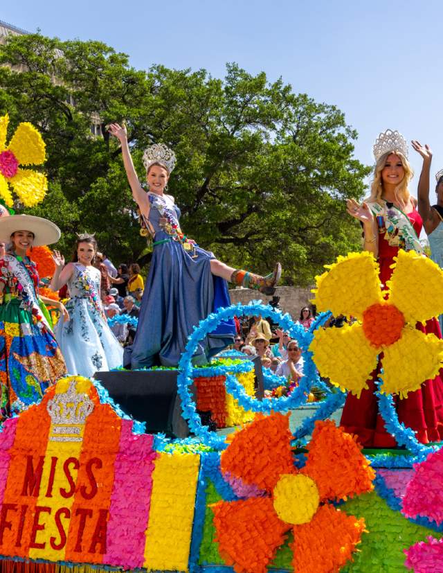 Women dressed in gowns and crowns on a colorful Fiesta float in front of the Alamo.