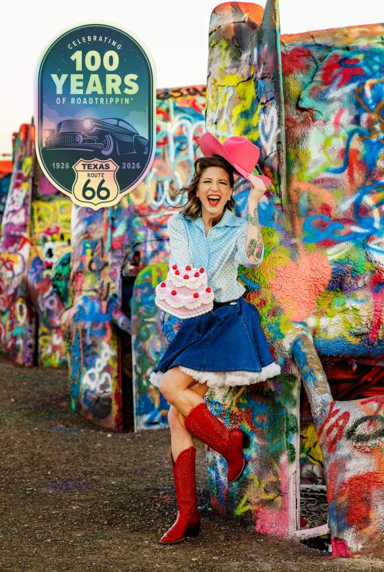a woman in a pink cowboy hat and red boots laughs while holding a small birthday cake at Cadillac Ranch, leaning against the brightly painted cars as a badge marks 100 years of Texas Route 66 and the spirit of road tripping in Amarillo