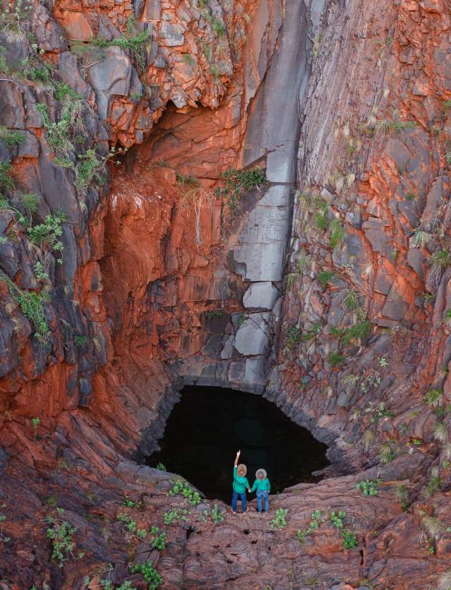 Two people stand at the base of a gorge with deep red walls at Cheela Plains in the Pilbara. There is a pool of water at the bottom, and marks on the gorge walls indicating that a waterfall flows when it rains.
