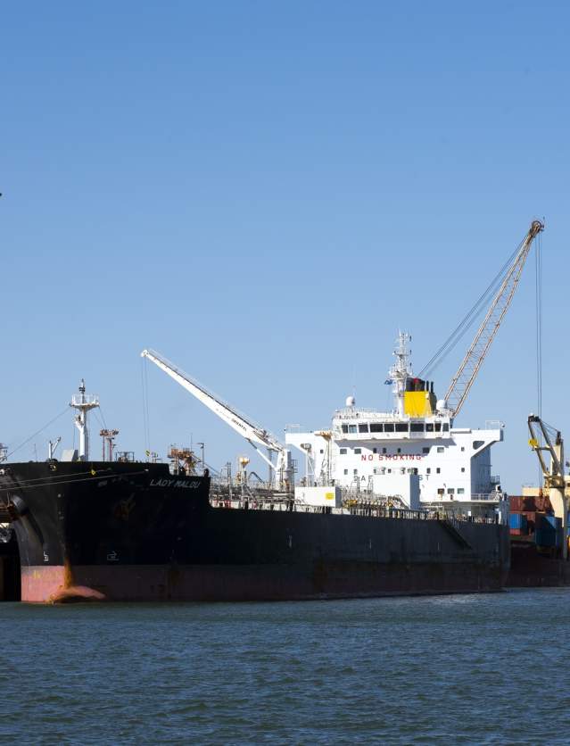 An Iron ore carrier in the Port of Port Hedland, seen from the water with a tug boat along side and a crane on land.