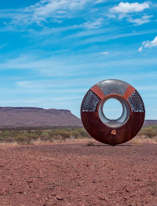 A view of the Resilience Sculpture outside Paraburdoo with mountains in the background. A pair of boots hangs off the sculpture