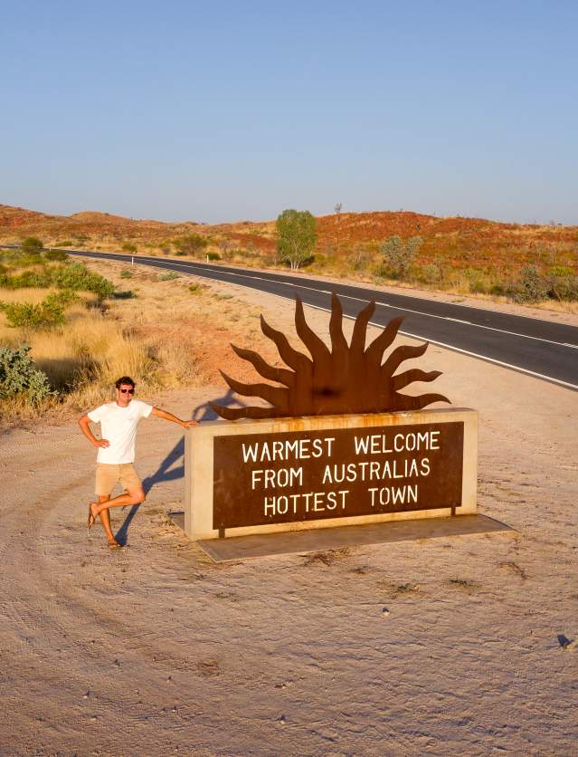 Man standing at the Marble Bar welcome sign which reads "Warmest welcome from Australia's hottest town."