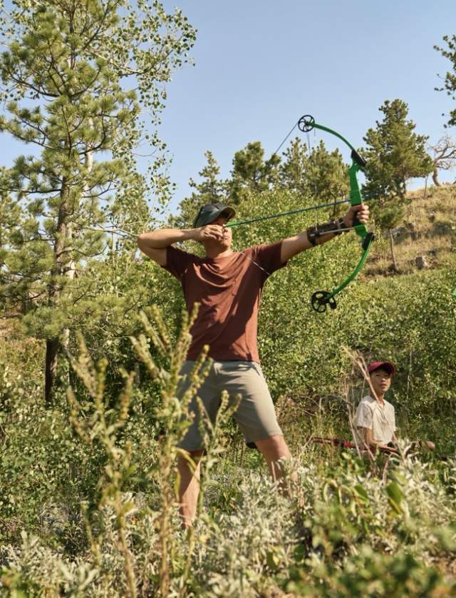 man holding bow and arrow pointed toward target with child and guide as audience