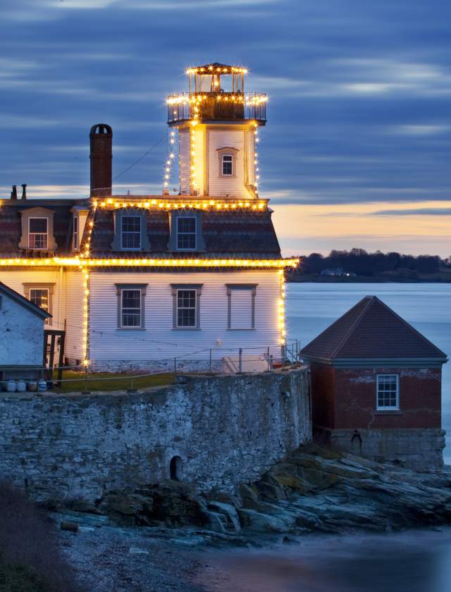 A white building with a lighthouse on top, decorated with lights, sits on a rocky waterfront at dusk.