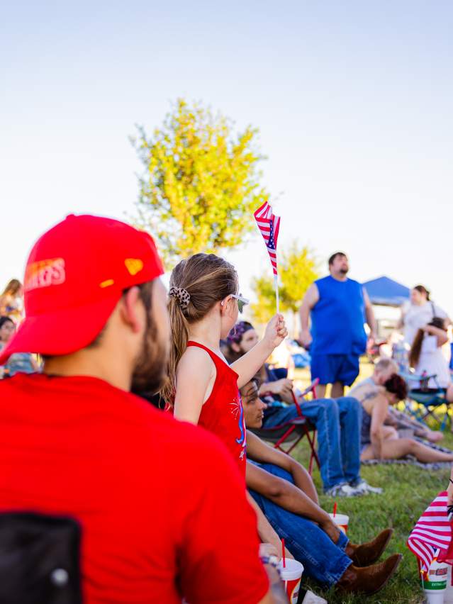 A crowd at a 4th of July event with a girl in a USA headband and star sunglasses.
