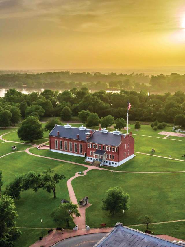 The sun is shining behind clouds above the National Historic Site in Fort Smith, Arkansas with the Arkansas River flowing behind it.